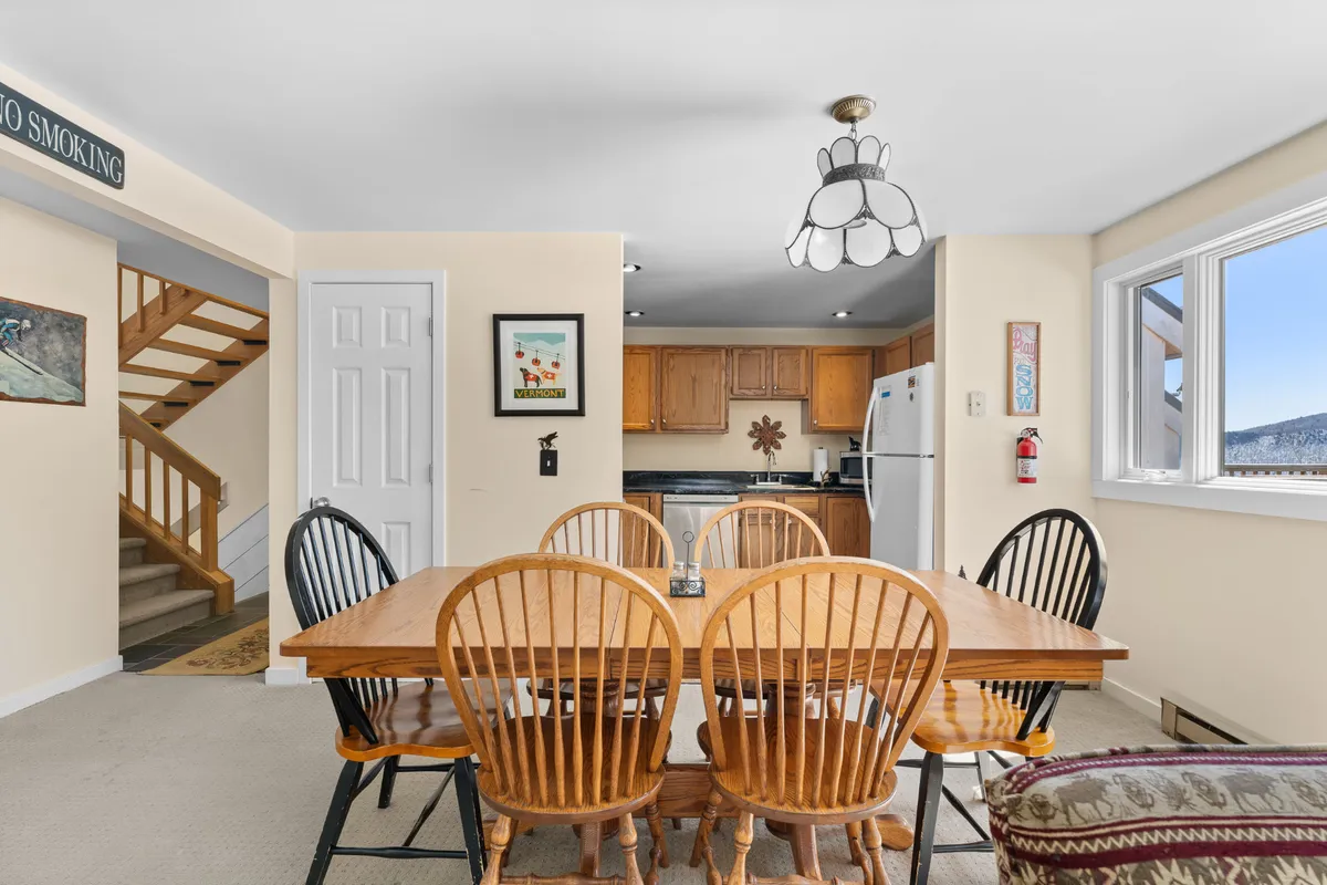 A cozy dining area featuring a wooden dining table, visible kitchen area, staircase, and a window with a view. The setting feels warm and communal, perfect for intimate family dinners, socializing, or enjoying sunrise breakfasts together.