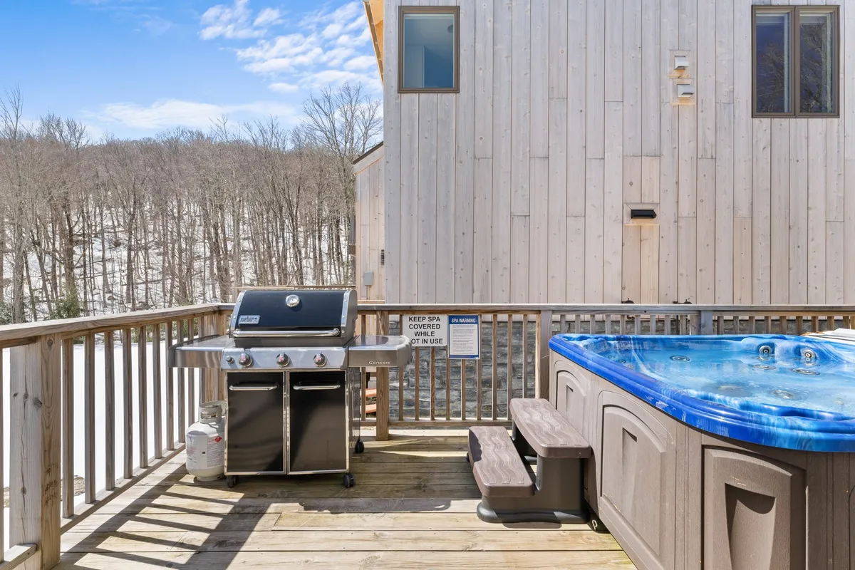 An inviting exterior deck scene featuring a grill, hot tub, and surrounding trees. The space is open to the outdoors, perfect for relaxing with your pet or unwinding after a day of hiking or snowshoeing. The wooded backdrop hints at immediate access to nature.