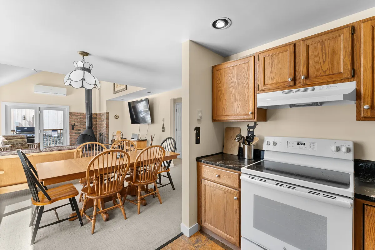 A warm, open-plan dining space with a wooden dining table in the foreground, modern kitchen appliances and cabinetry in the background, and a soft light fixture overhead. The scene feels inviting and set for family meals or gatherings, with the kitchen and dining areas seamlessly connected.
