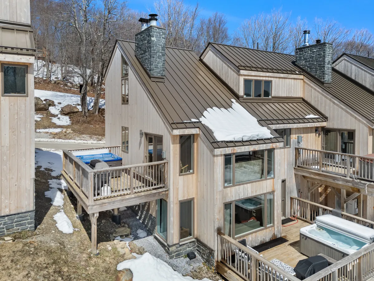 A view of the home’s exterior deck, showing a hot tub, large windows, and a wooden exterior with a stone chimney. The setting hints at the option for deck dining and outdoor gatherings, with the promise of memorable meals enjoyed against a scenic Vermont backdrop.