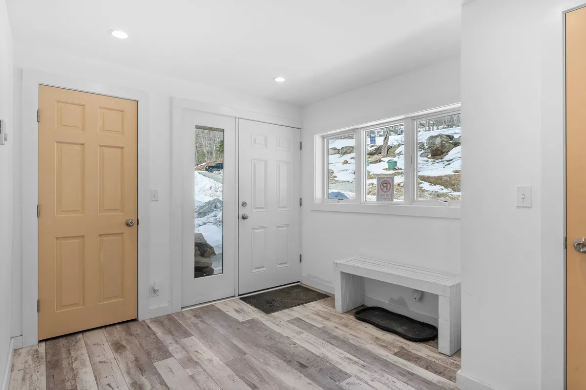 This image captures an inviting entryway featuring two doors (one white, one yellow), a white bench for removing shoes, and a large window with views of the snow-covered ground. The natural light highlights the functionality of the space and suggests an organized area for arrivals and departures, perfect for stashing outdoor gear.