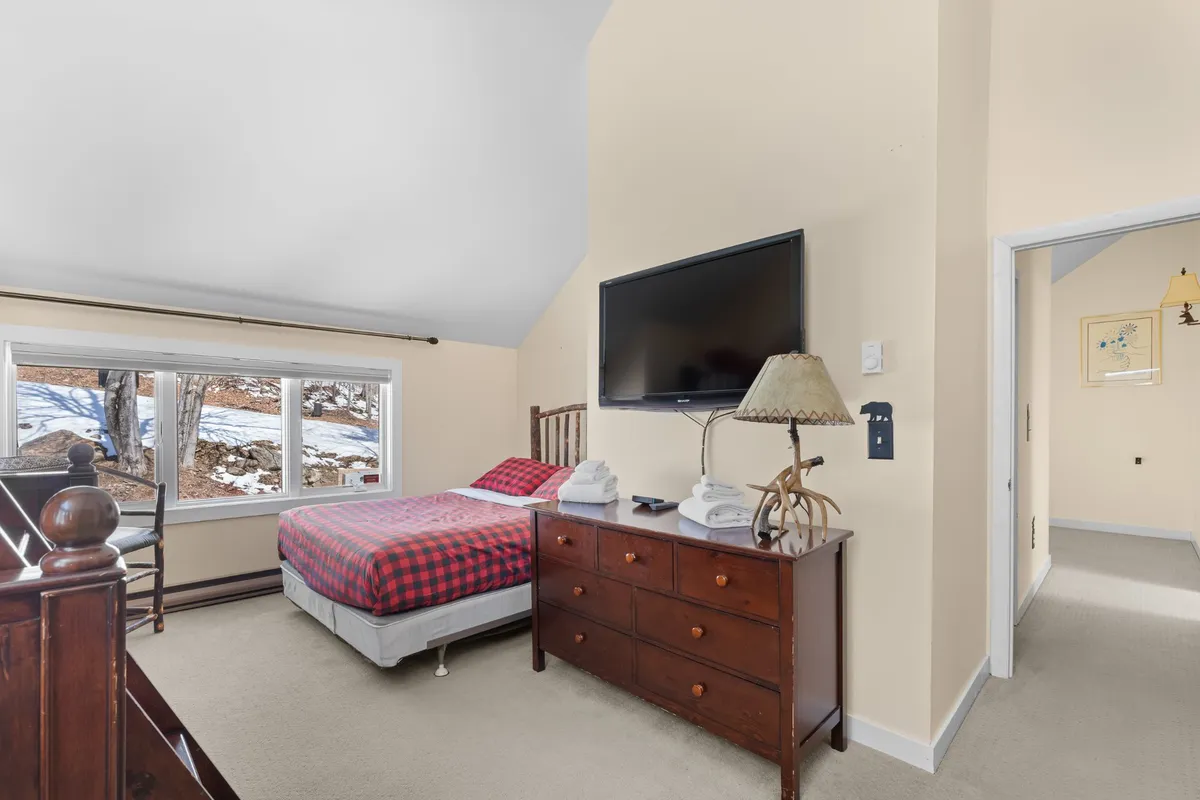 A cozy bedroom featuring a bed with plaid bedding, a wall-mounted television, and a wooden dresser. Natural light fills the space, and there is a comfortable, welcoming ambiance that invites relaxation and restful sleep.