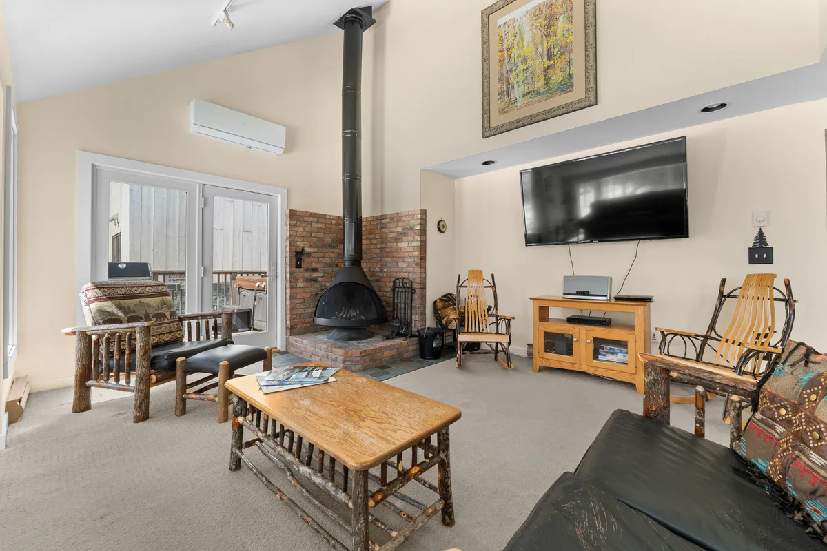 A cozy living room featuring a wood-burning stove, comfortable seating with plush cushions, a rustic coffee table, and a television. Large windows let in ample natural light and offer glimpses of the surrounding landscape, creating a warm and inviting atmosphere ideal for relaxation after a day outdoors.