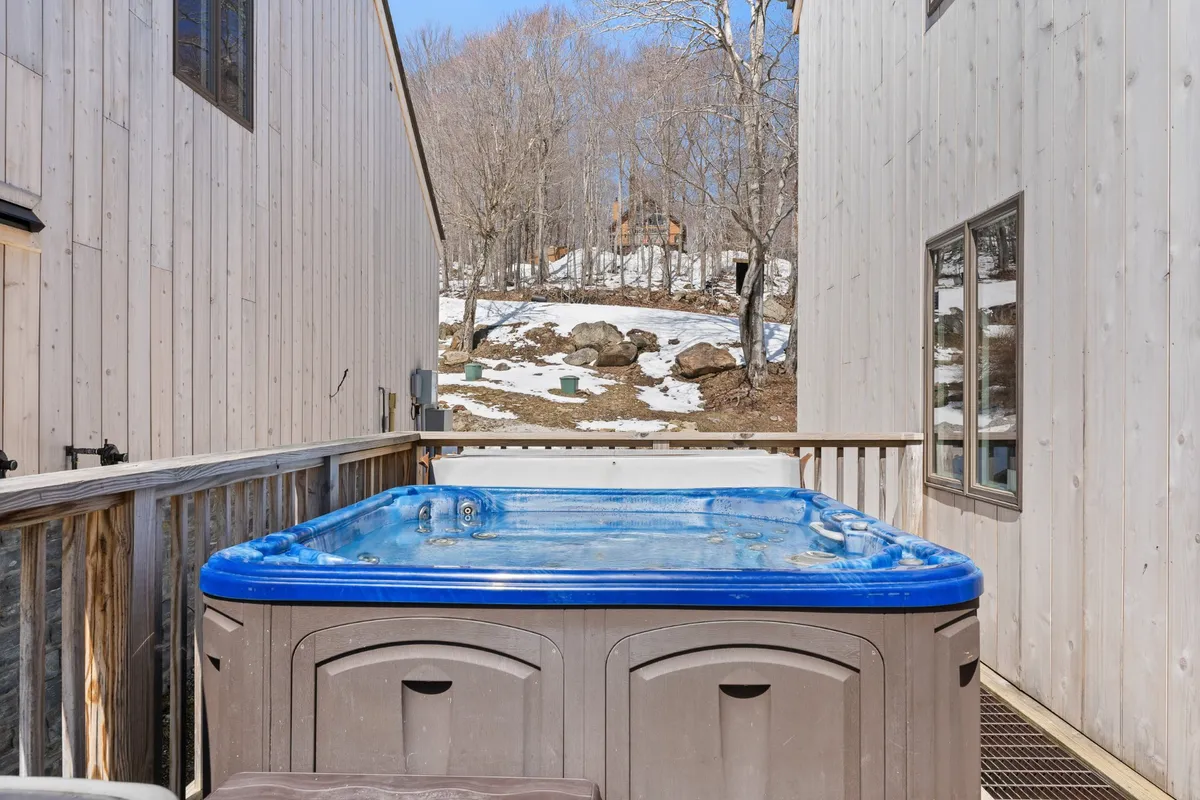 An exterior view of a wooden deck with a hot tub surrounded by lush trees. The setting is tranquil, with the deck providing space for outdoor gatherings or relaxing soaks while taking in natural Vermont scenery.