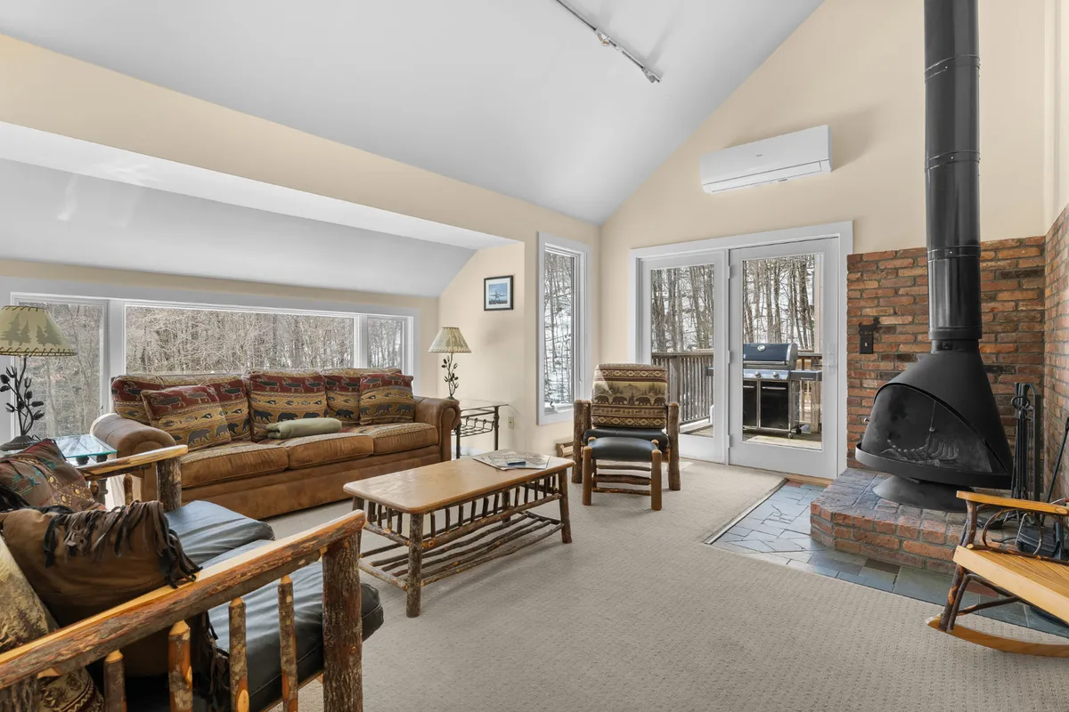 The image shows a warm, inviting living room inside a slopeside condo. Centered around a glowing fireplace, a plush sofa and wooden chairs offer cozy spaces to relax. Large windows frame snowy forest views, filling the space with natural light and a sense of connection to the outdoors.
