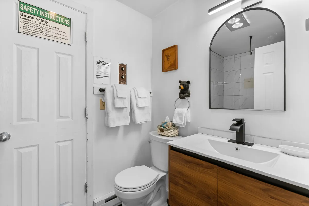 A bright, inviting bathroom scene featuring a modern sink, large mirror, wooden cabinetry, and a sign with safety instructions. The space feels functional and well-maintained, ideal for post-sauna or hot tub wash-up and self-care routines.