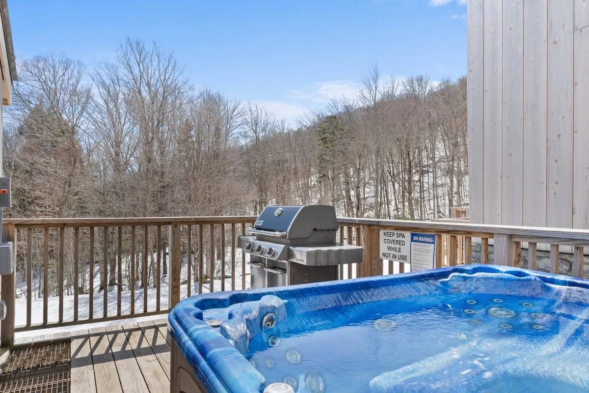This exterior image features a wooden deck surrounded by trees, complete with a hot tub and barbecue grill. The setup invites guests to relax outdoors, enjoy a soak, and share meals in a natural setting. The deck is spacious, making it ideal for group gatherings or family BBQs.